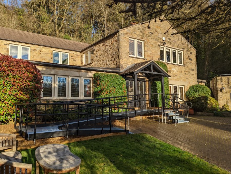 A wheelchair ramp going up and back on itself on the left handside of a stone effect home. While featuring steps up onto the top platform which continues up to the entrance doors.