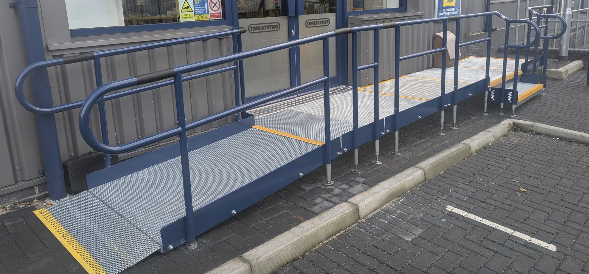 Image of a metal modular ramp with blue handrails. The ramp has a yellow stripe accross the bottom of the ramp to outline where the bottom of it is. It has a step unit toward the right middle of the ramp. It is outside a Toolstation shop in Portsmouth.