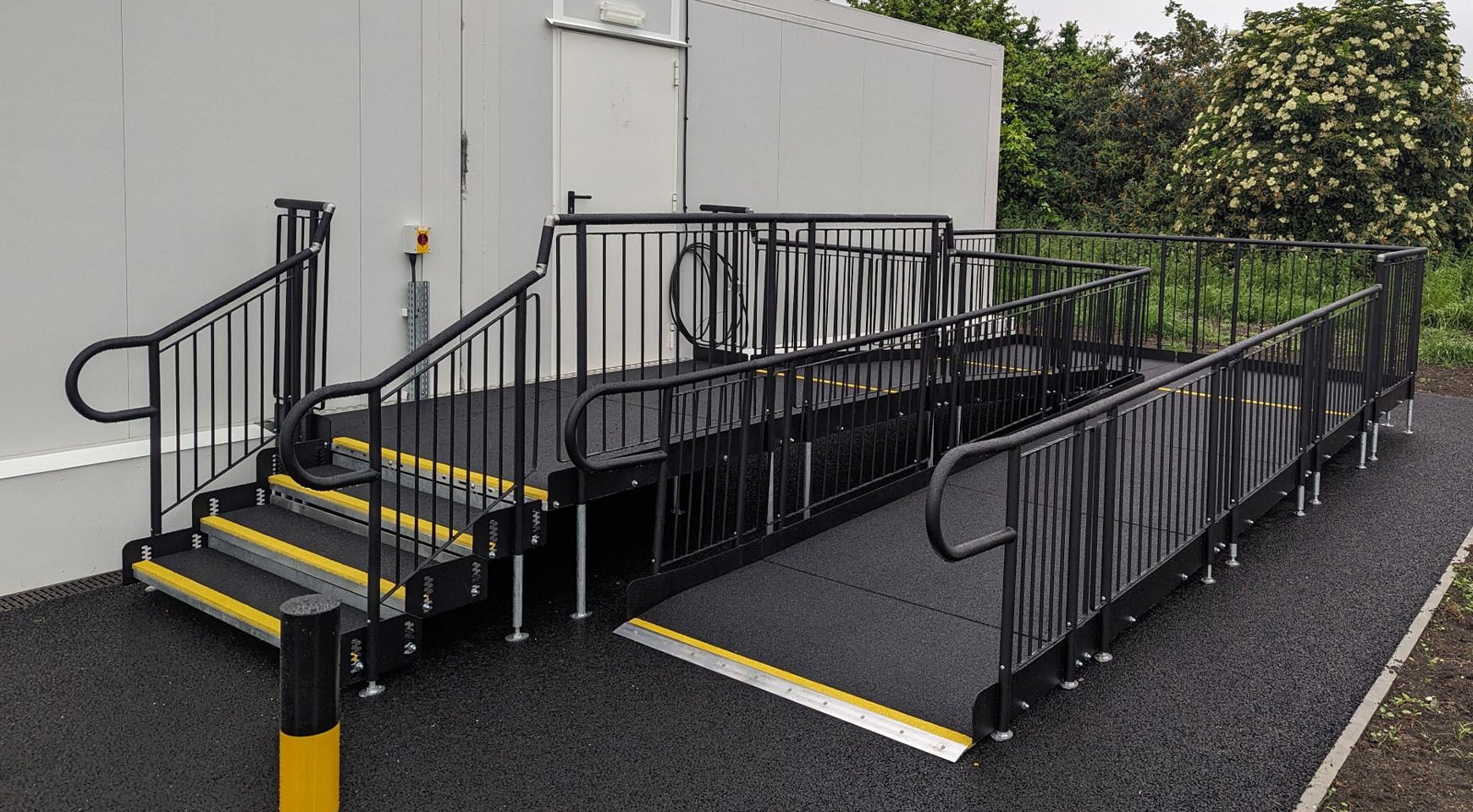 A black metal modular ramp with handrails going veritcal upright. It also has a step unit for ambulant users. This ramp and step unit is leading up towards a door.