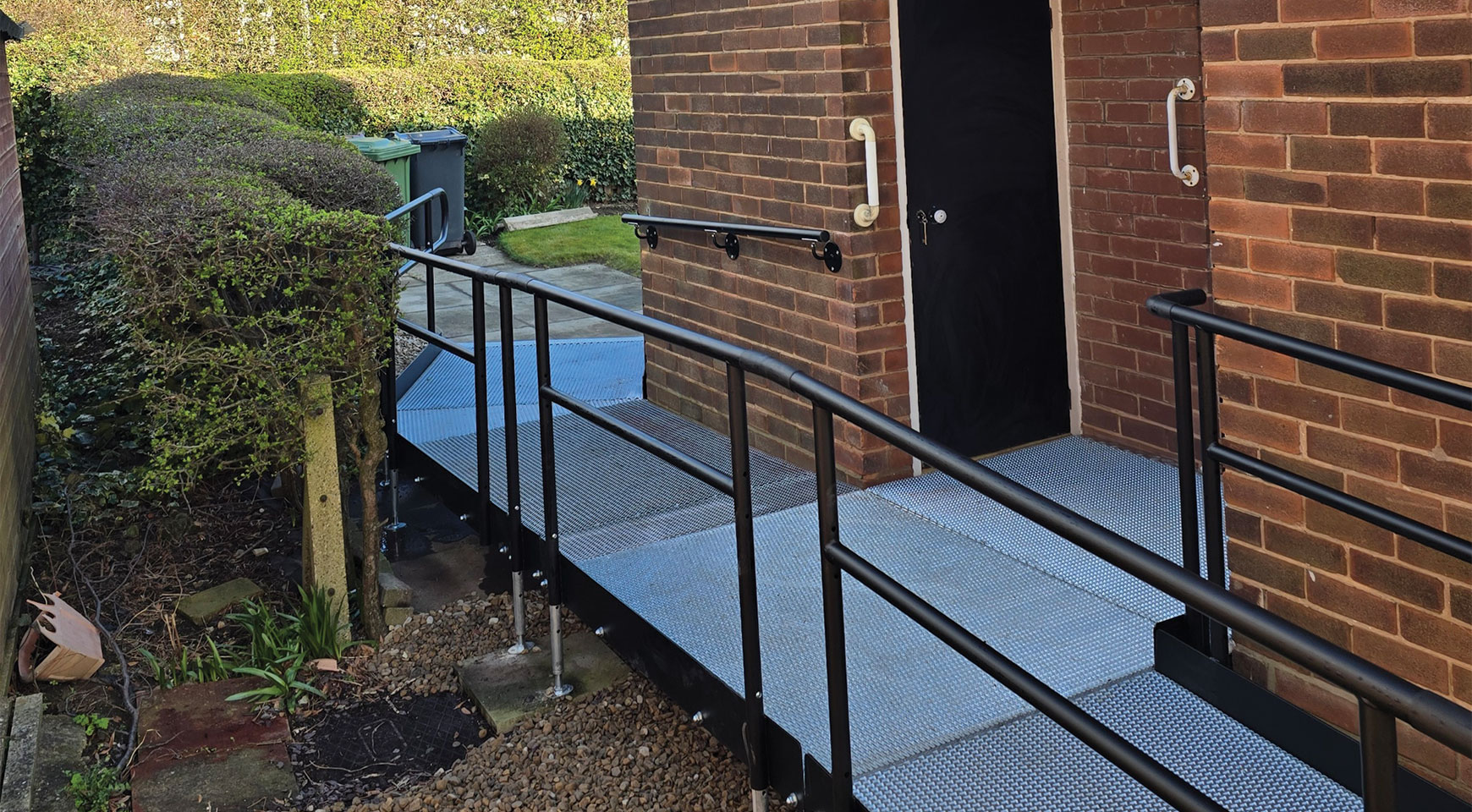 The image shows a metal accessibility ramp leading to a brick building entrance. It features sturdy handrails, a non-slip surface, and a gentle slope suitable for wheelchairs. Grab handles are mounted near the door, and the area is clean and well-maintained.