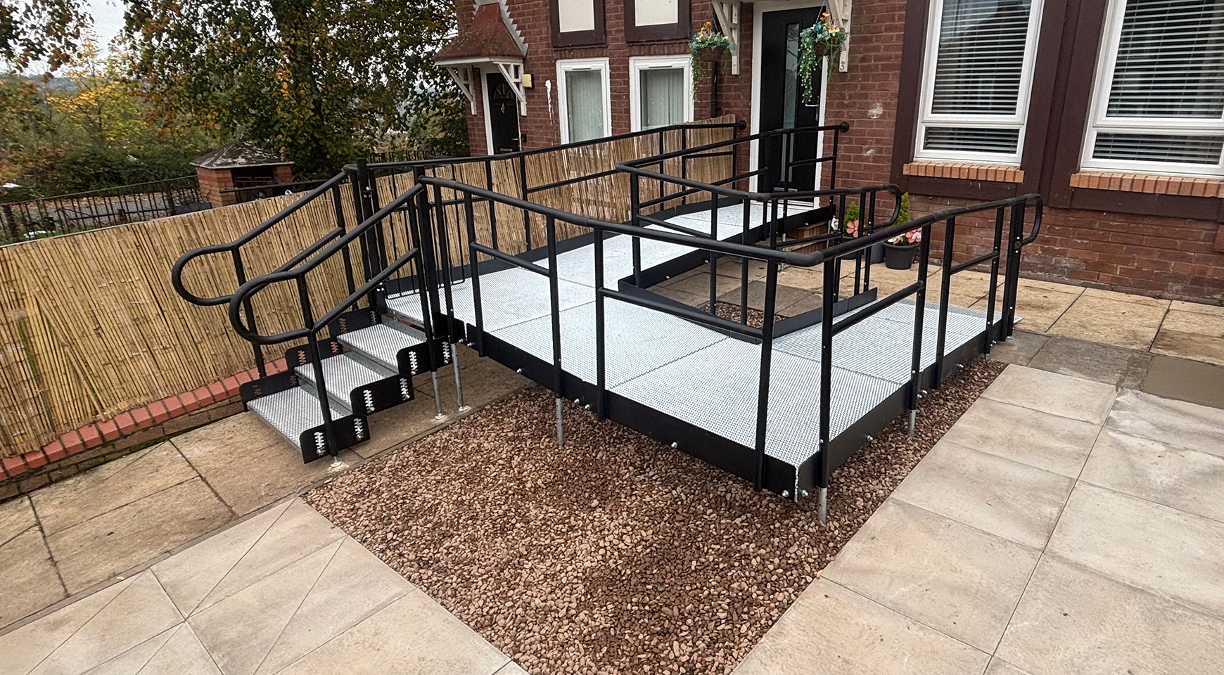 A black bespoke modular access ramp with handrails and a three tread step unit coming off the side, installed at the front entrance of a brick residential property. Its designed to overcome a sloped garden and provide safe, level access to the front door.