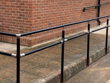 Black metal handrail system with integrated midrail installed along a gently sloped pavement beside a brick building.