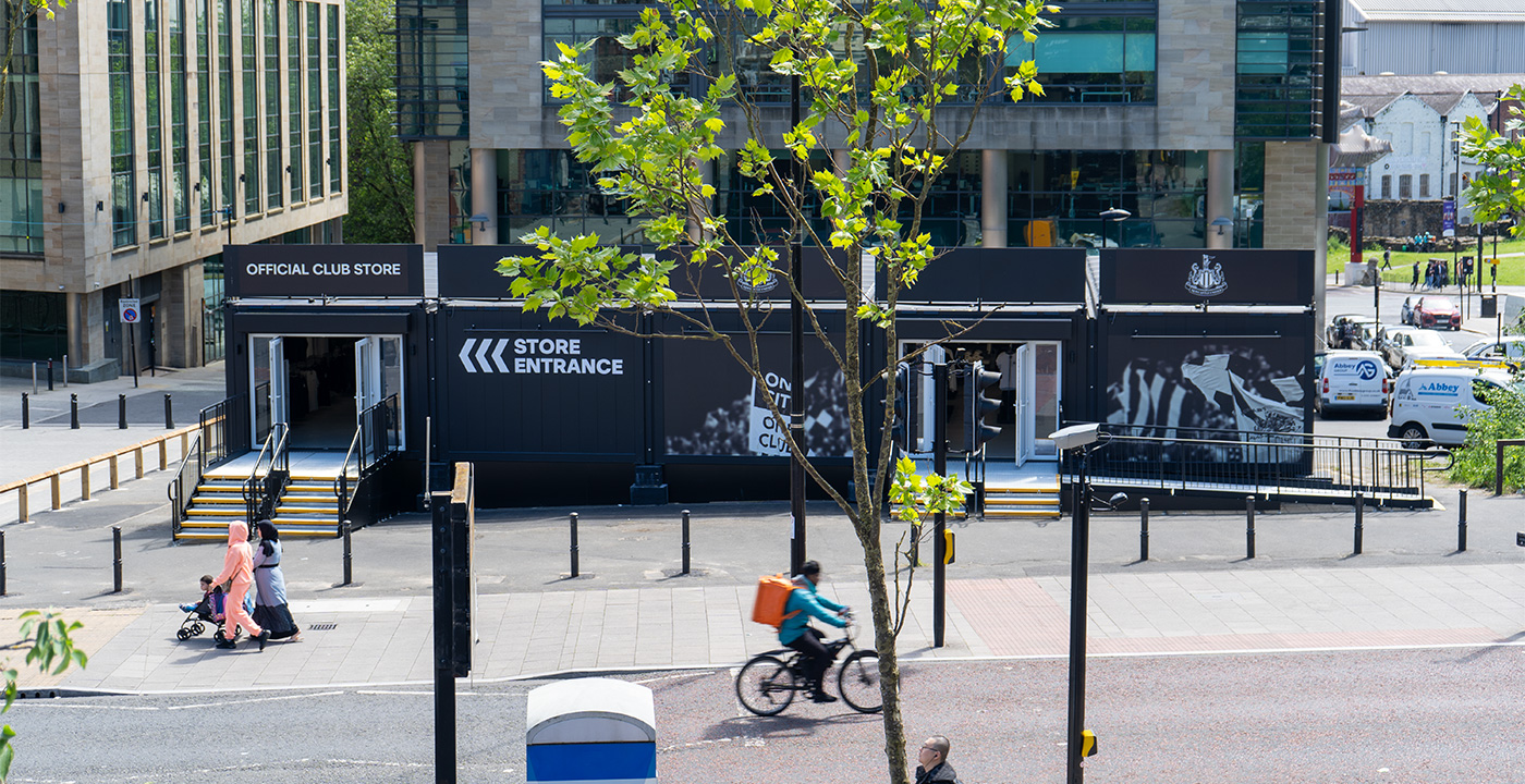 A black modular building with newcastle United Football Club Store just off of a main road. A double step unit is on the left to enter the shop. There is a modular ramp on the right hand side that leads up to the shop, with a step unit working in conjunction. All steps and ramps have black hand rails, with a silver metal mesh surface.
