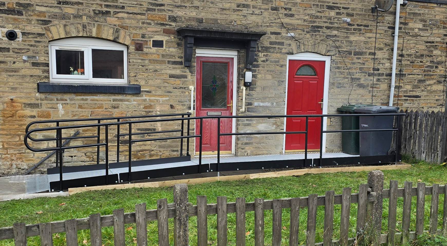 A sandstone brick house with a metal modular ramp infront of it. There are 2 red doors on the house and both lead out to a platform. To the left of the platforms the ramp goes down to ground level. The ramp has black handrails and a silver surface. There is a window to the left of the red doors.
