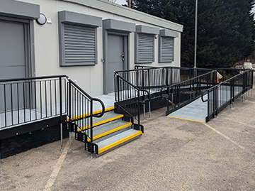 A white modular building with a black railed modular ramp infront of it. The ramp also had a step unit with yellow nosing on each step. The steps lead straight to the modular building whereas the ramp goes vertical along the building and turns back on itself for access to the building.