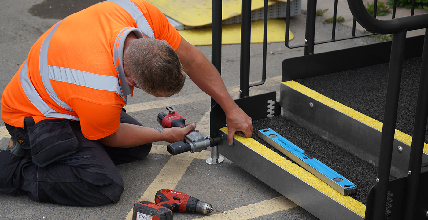 Installer securing a modular ramp step unit in place using power tools, with a spirit level positioned on the step for accuracy.