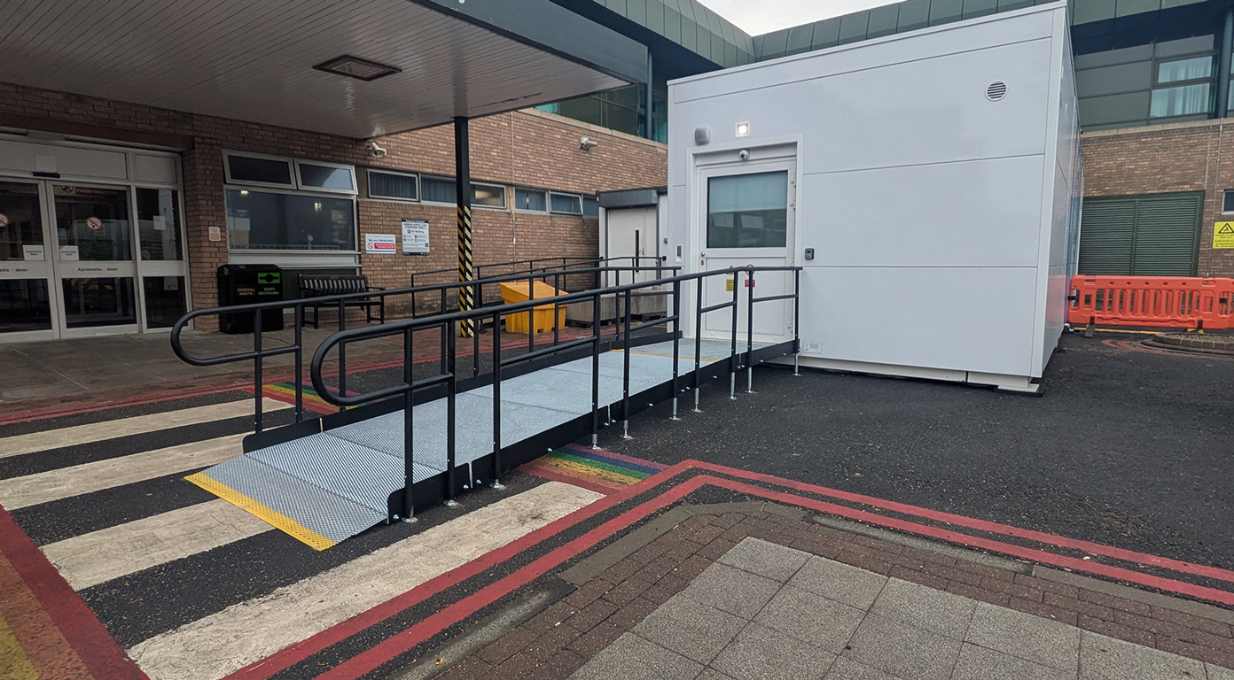 A modular ramp leading to a white modular building outside a hospital. The rails are black with a silver metal mesh surface. There is a yellow strip at the start of the ramp which is yellow nosing as a visual cue that the ramp is starting.